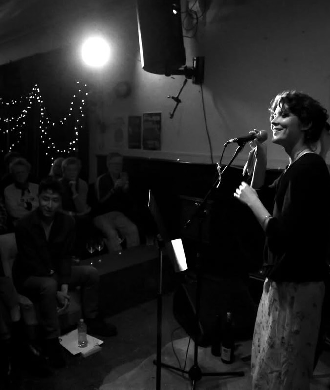 Eartha Davis singing into a microphone at an intimate live performance, framed by fairy lights